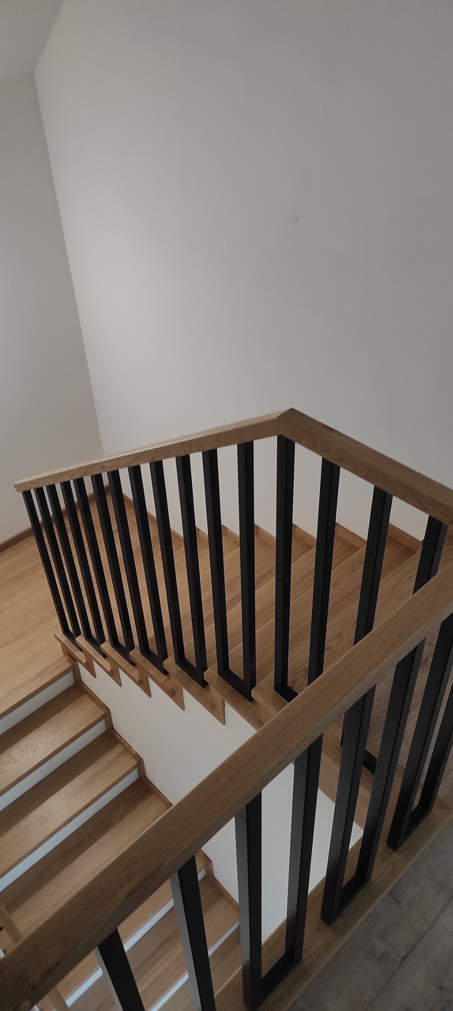 Nowoczesne schody z drewnianymi stopniami i czarną, metalową balustradą, widok z góry na jasne wnętrze.