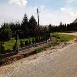 Nowo postawione, ciemne ogrodzenie z paneli siatkowych na betonowej podmurówce, oddzielające posesję od wiejskiej drogi; w tle wysokie drzewa iglaste i błękitne niebo z chmurami.