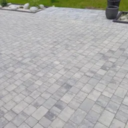 Ułożona szara kostka brukowa na podjeździe, z dekoracyjnymi kamieniami i roślinami wzdłuż krawędzi, oraz elementami wyposażenia ogrodu w tle.