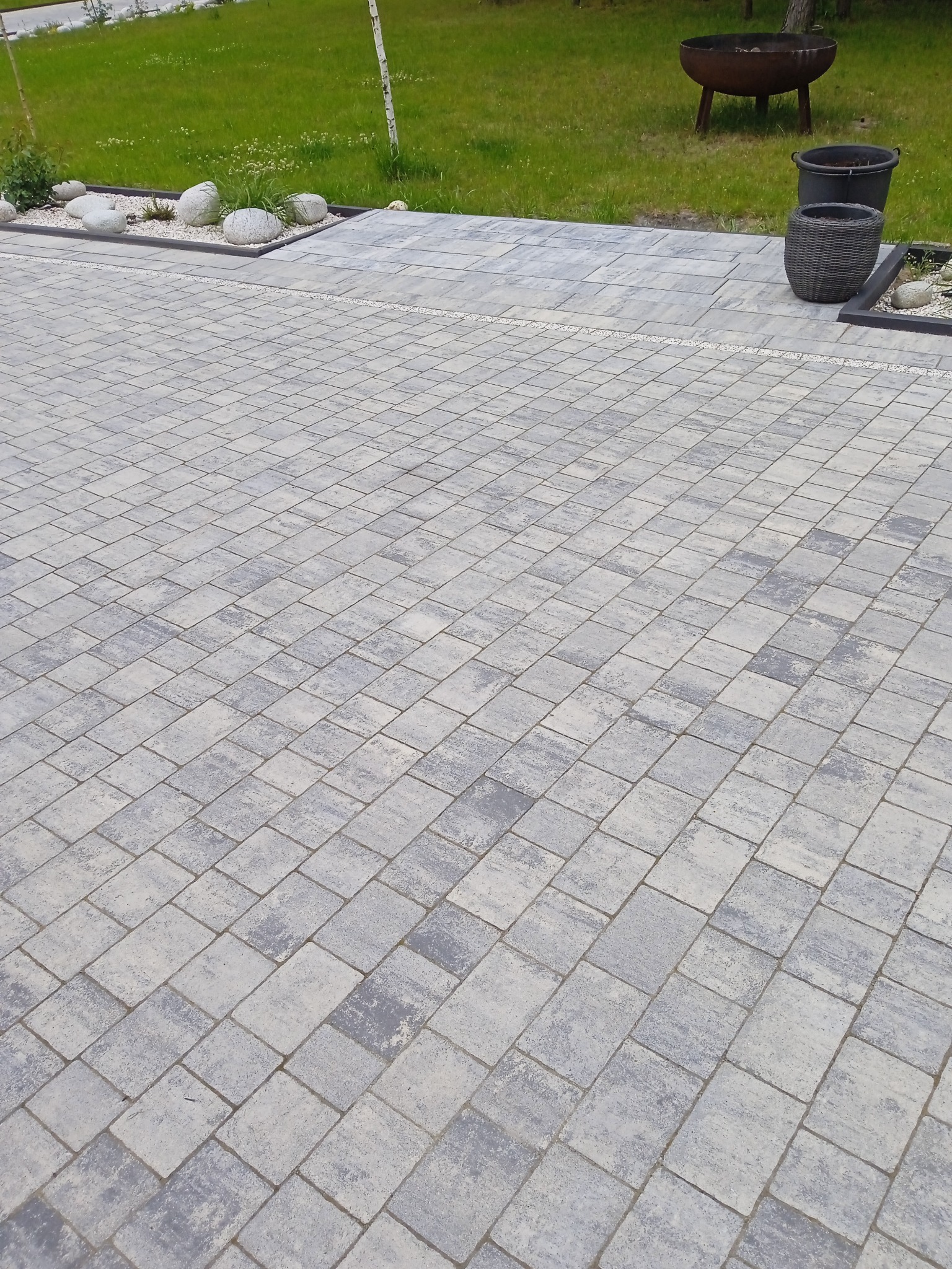 Ułożona szara kostka brukowa na podjeździe, z dekoracyjnymi kamieniami i roślinami wzdłuż krawędzi, oraz elementami wyposażenia ogrodu w tle.