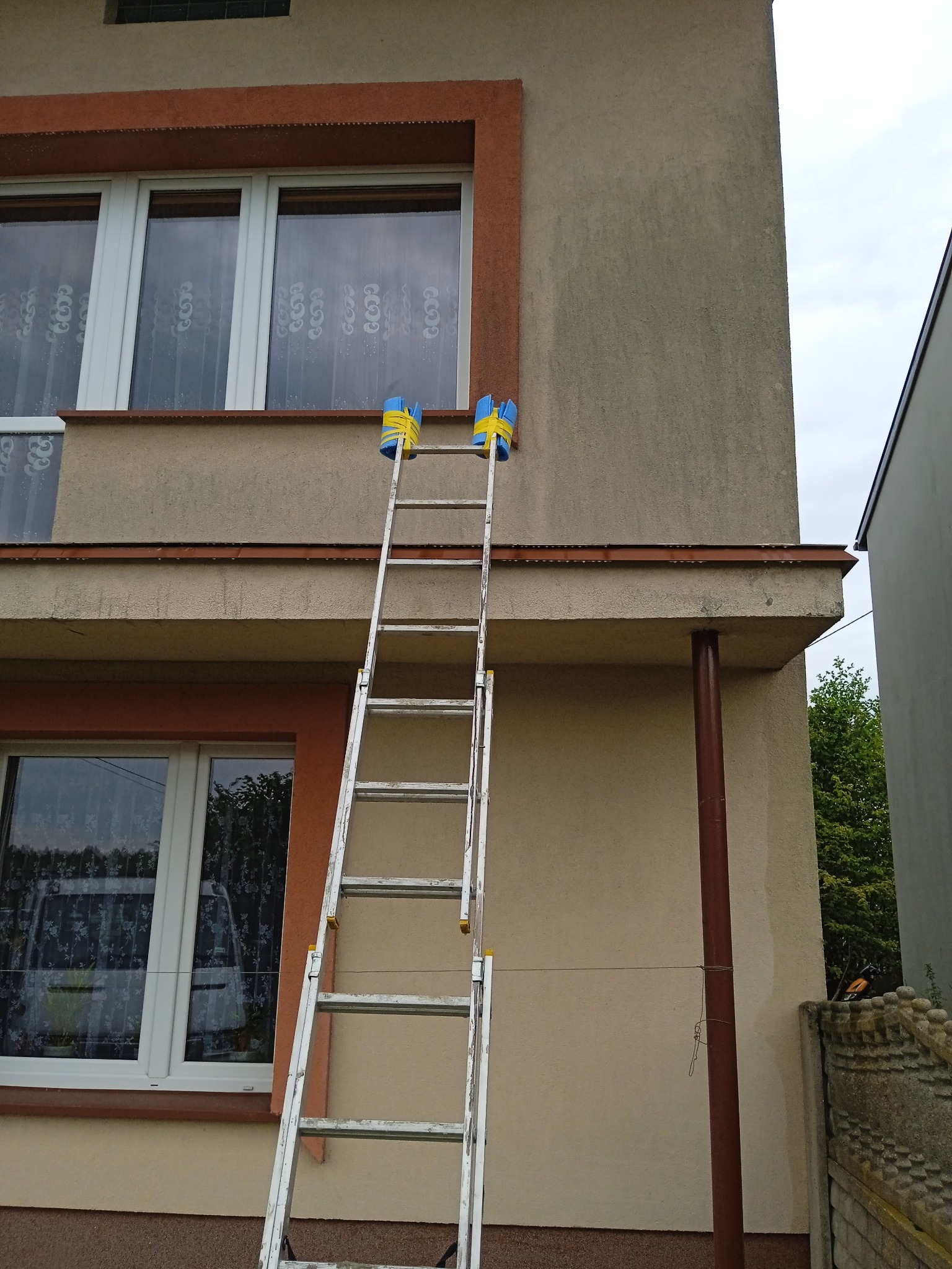 Aluminiowa drabina oparta o elewację budynku, z dwoma wiaderkami zawieszonymi na górnych szczeblach, przygotowana do prac malarskich lub czyszczenia.