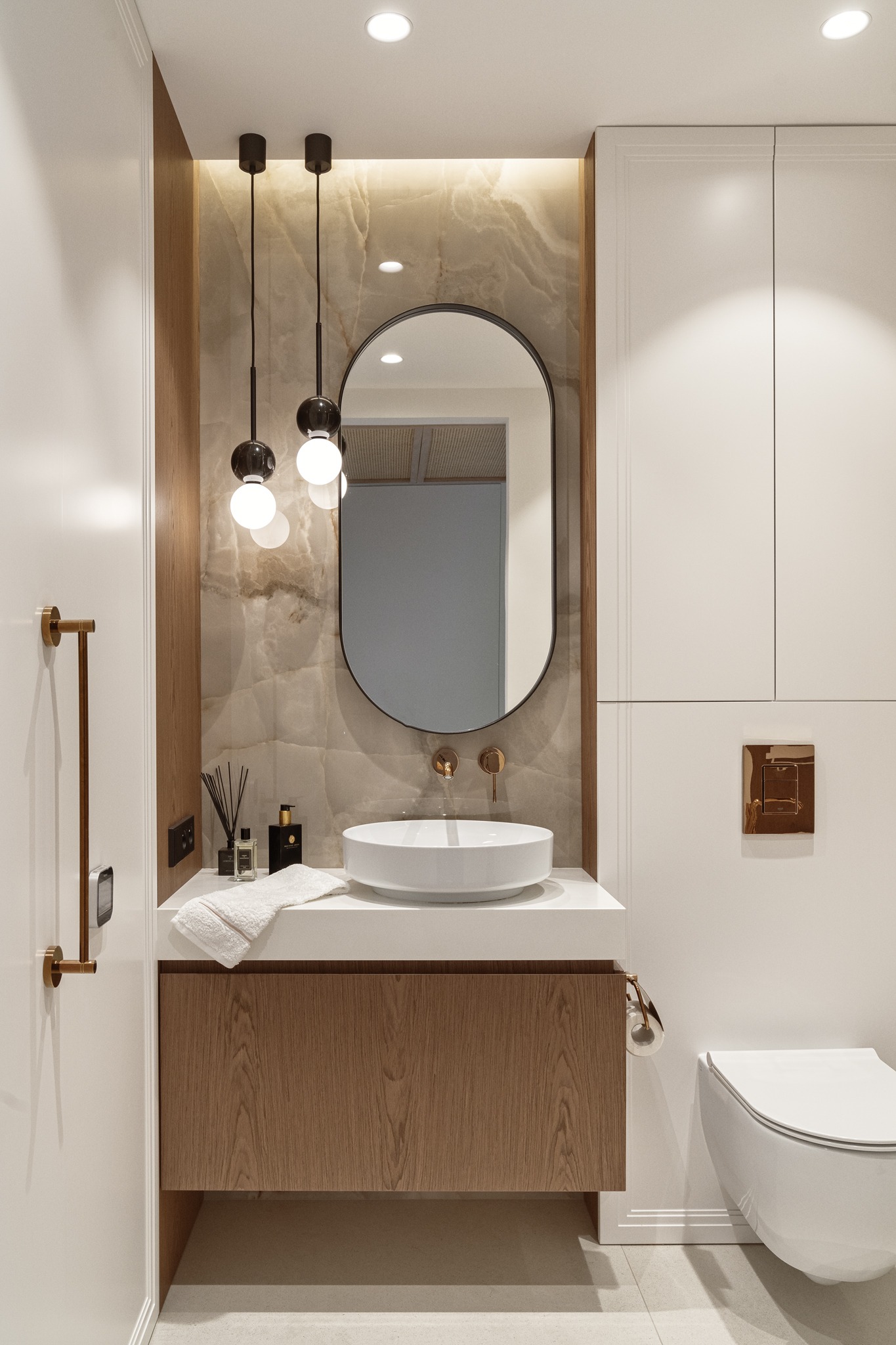 Nowoczesna łazienka z owalnym lustrem, marmurową ścianą, wiszącą umywalką, drewnianą szafką i złotymi akcentami, oświetlona wiszącymi lampami kulami.