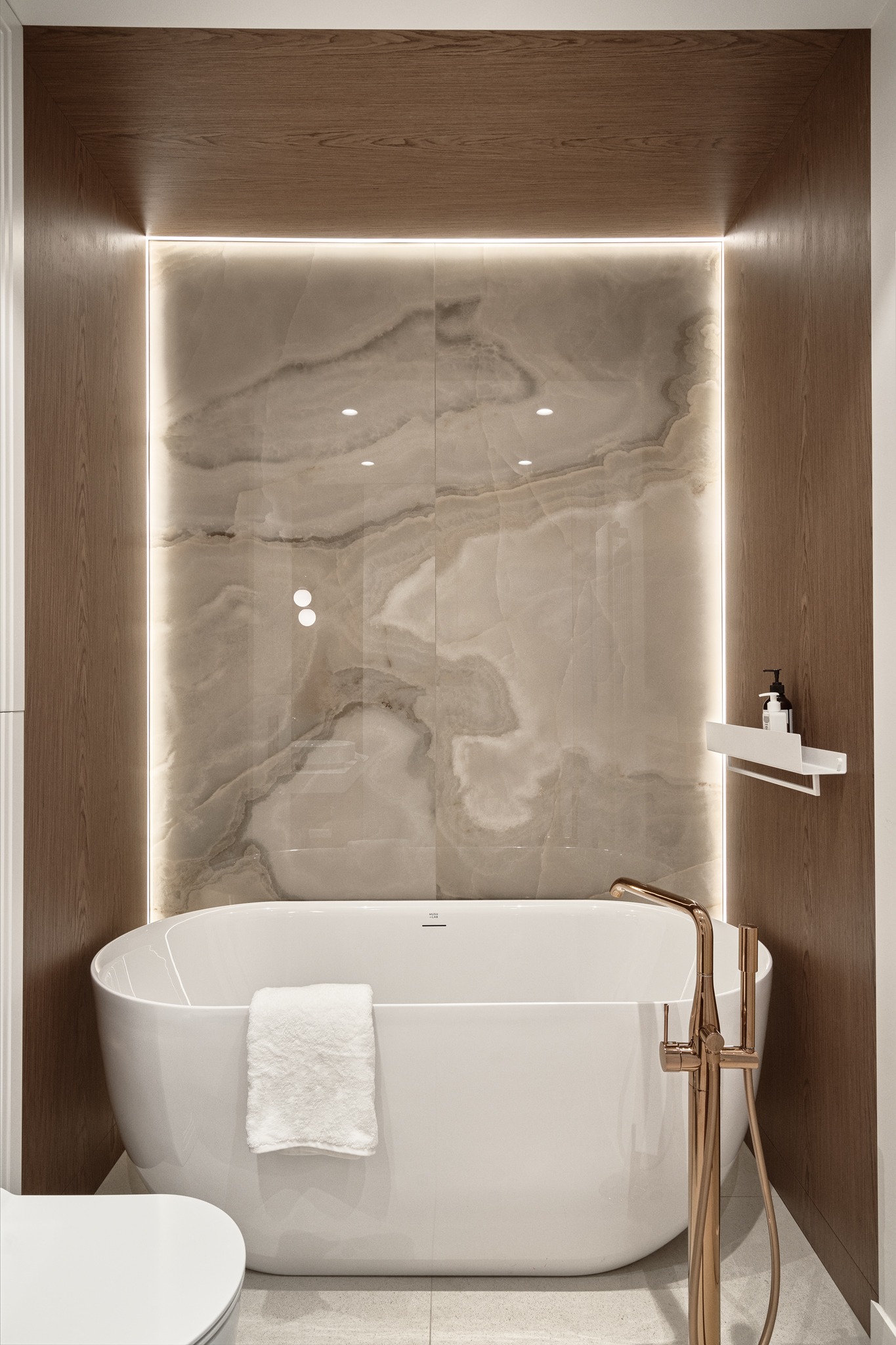 Nowoczesna łazienka z wolnostojącą białą wanną, marmurową ścianą z podświetleniem LED i złotą armaturą.