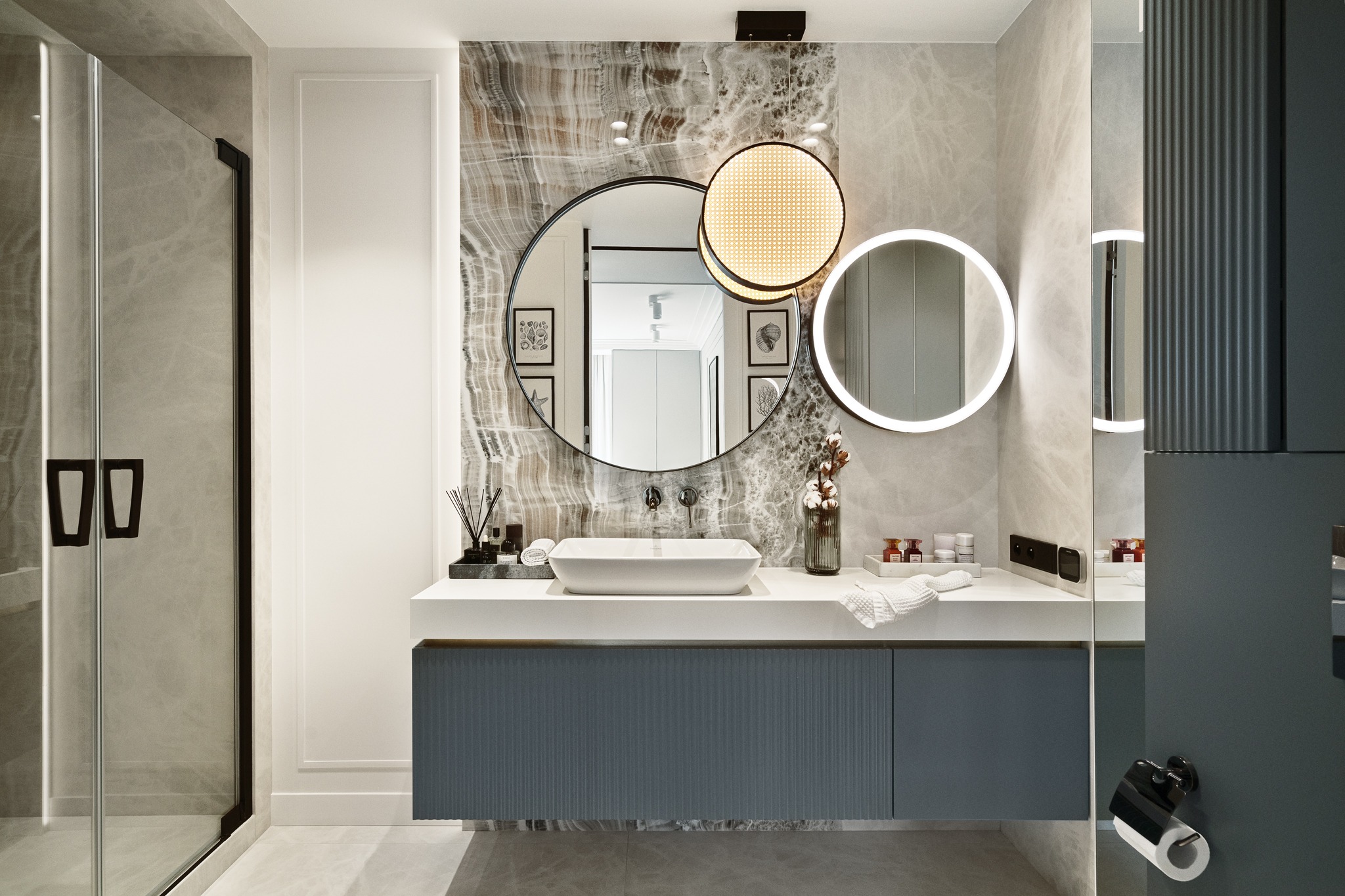 Elegancka łazienka z marmurową ścianą, okrągłymi lustrami z podświetleniem LED, wiszącą szafką podumywalkową w kolorze grafitowym z ryflowanym frontem i kabiną prysznicową ze szklanymi drzwiami.
