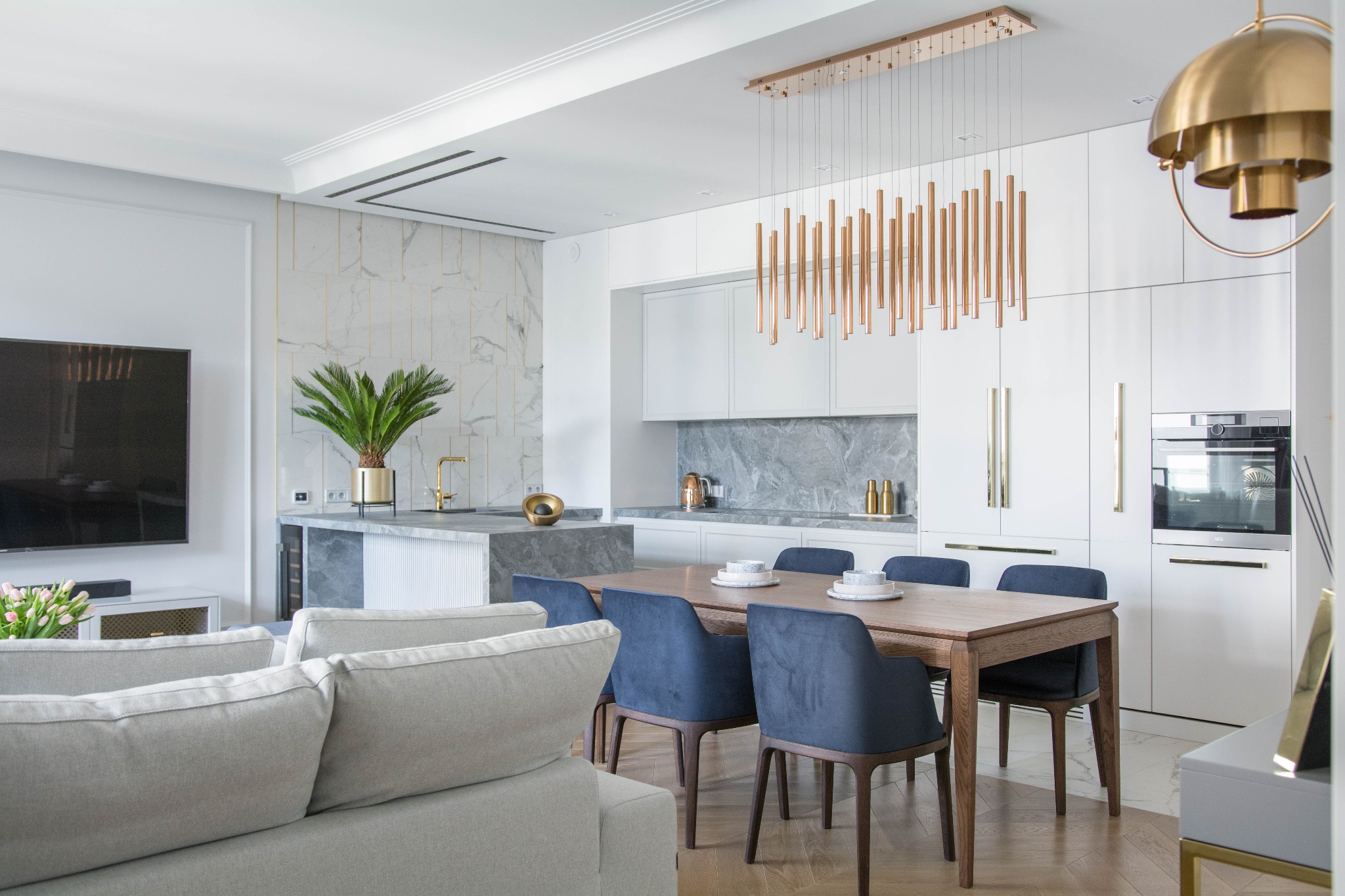 Jasne wnętrze: salon z kuchnią, drewniany stół z niebieskimi krzesłami, marmurowa ściana, złote akcenty i nowoczesne oświetlenie.
