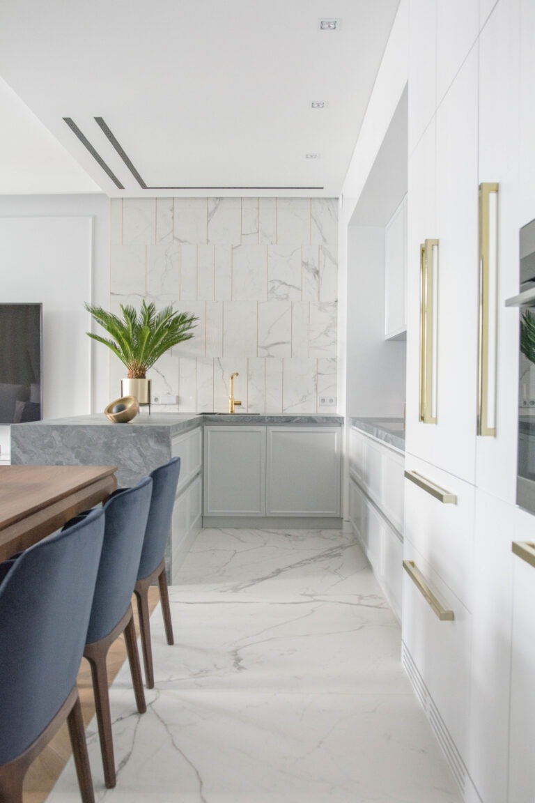 Jasna kuchnia z marmurową ścianą akcentową z delikatnymi złotymi wstawkami, szarą wyspą z blatem z kamienia, złotą baterią i rośliną doniczkową. Widoczne krzesła z niebieską tapicerką i drewnianymi...
