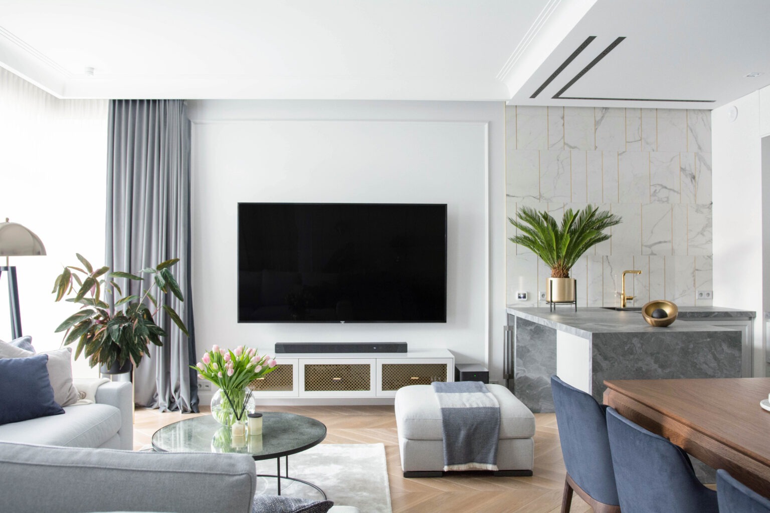 Jasne, przestronne wnętrze salonu z aneksem kuchennym w stylu nowoczesnym, z telewizorem na ścianie, szarymi zasłonami i parkietem w jodełkę.
