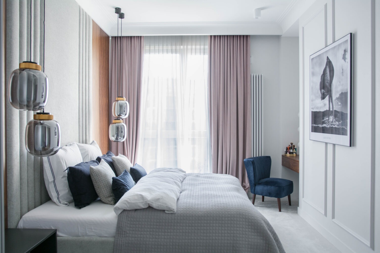 Elegancka sypialnia z tapicerowanym panelem ściennym za łóżkiem, wiszącymi szklanymi lampami i zasłonami w odcieniach różu, uzupełniona o granatowe poduszki i krzesło.