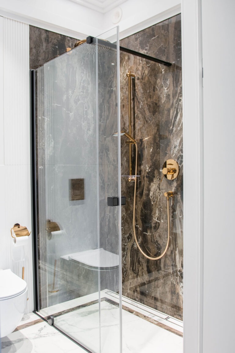 Nowoczesna kabina prysznicowa typu walk-in z marmurowymi ścianami, złotą armaturą i odpływem liniowym, widok z zewnątrz na toaletę z elementami w kolorze złota.