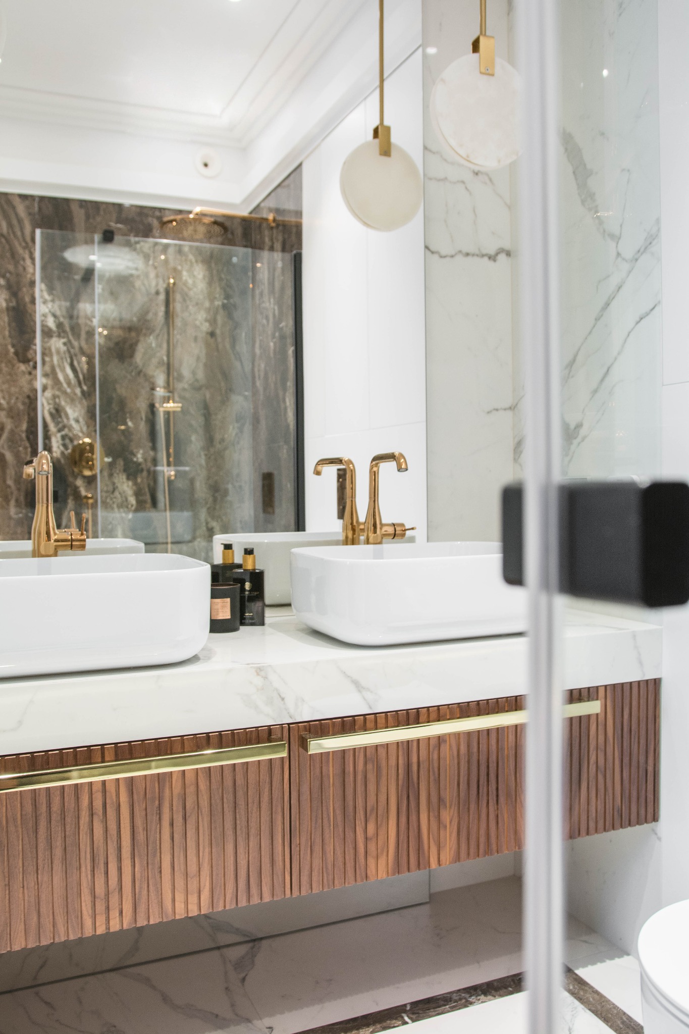 Elegancka łazienka z dwoma białymi umywalkami nablatowymi, złotymi bateriami, marmurowym blatem i szafką z ryflowanymi frontami, widoczna kabina prysznicowa z marmurową ścianą w tle.