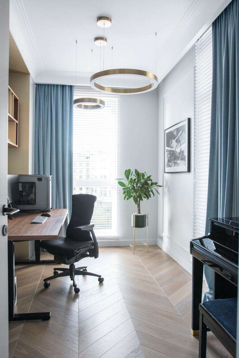 Jasne biuro z parkietem w jodełkę, biurkiem z komputerem, czarnym krzesłem obrotowym, pianinem i rośliną w złotej donicy, oświetlone dwoma okrągłymi lampami wiszącymi.