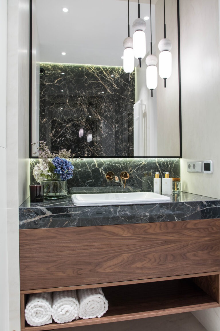 Nowoczesna łazienka w Warszawie z umywalką nablatową na blacie z czarnego marmuru, drewnianą szafką pod umywalką i okrągłymi lampami wiszącymi.