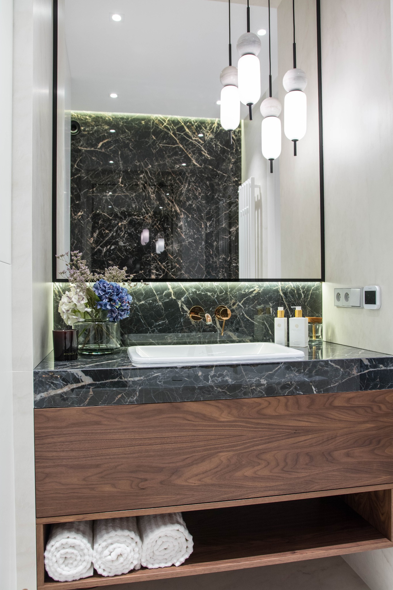 Elegancka łazienka z umywalką nablatową na blacie z czarnego marmuru, drewnianą szafką, złotą armaturą i dekoracyjnym oświetleniem punktowym odbijającym się w lustrze.