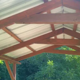 Wnętrze zadaszenia altany z widoczną konstrukcją drewnianą i blachą trapezową, z zielenią drzew w tle.