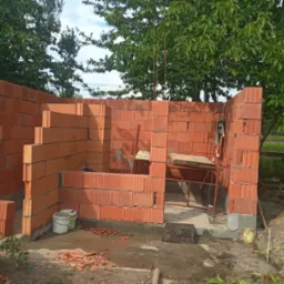 Wznoszenie ścian z czerwonej cegły dziurawki na fundamencie, widoczne rusztowanie, narzędzia i resztki zaprawy, zielone drzewa w tle.