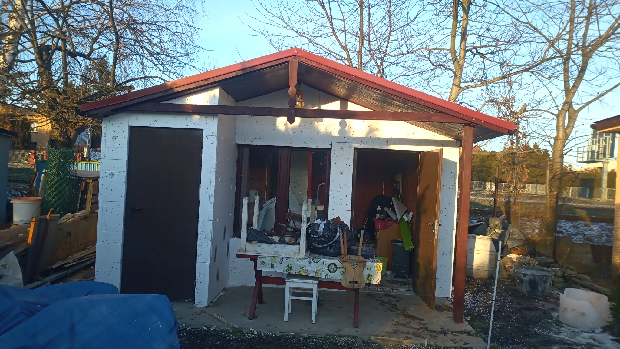 Niedokończony budynek gospodarczy z białych bloczków, z czerwonym dachem, widoczne drzwi, okno i otwarte wejście, przed budynkiem stół z krzesłem i bałagan