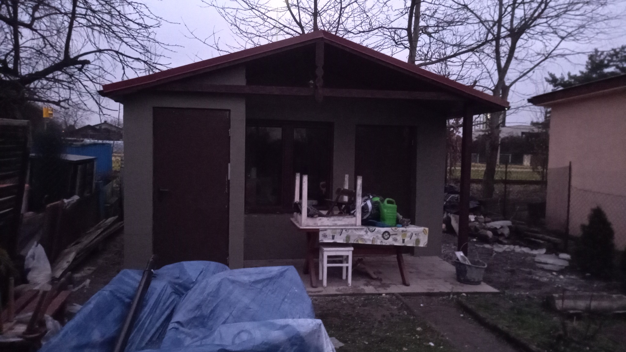 Szara altana z czerwonym dachem w ogrodzie, z widocznymi narzędziami i materiałami budowlanymi na zewnątrz, sugerującymi niedawne prace wykończeniowe.