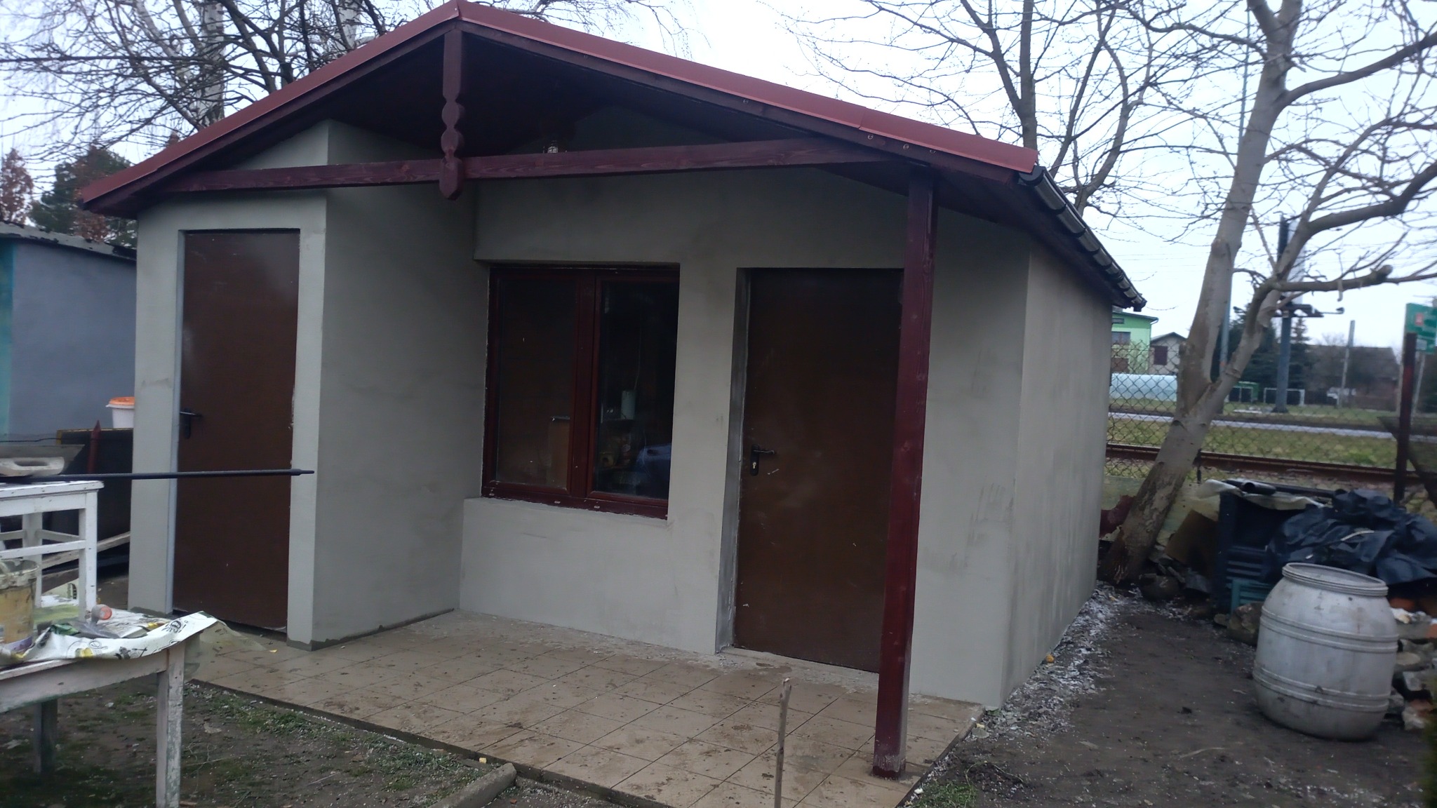 Nowo wybudowany, otynkowany budynek gospodarczy z dwoma brązowymi drzwiami i pojedynczym oknem, z czerwoną konstrukcją dachu, na zewnątrz widoczny stół roboczy z narzędziami i beczka.