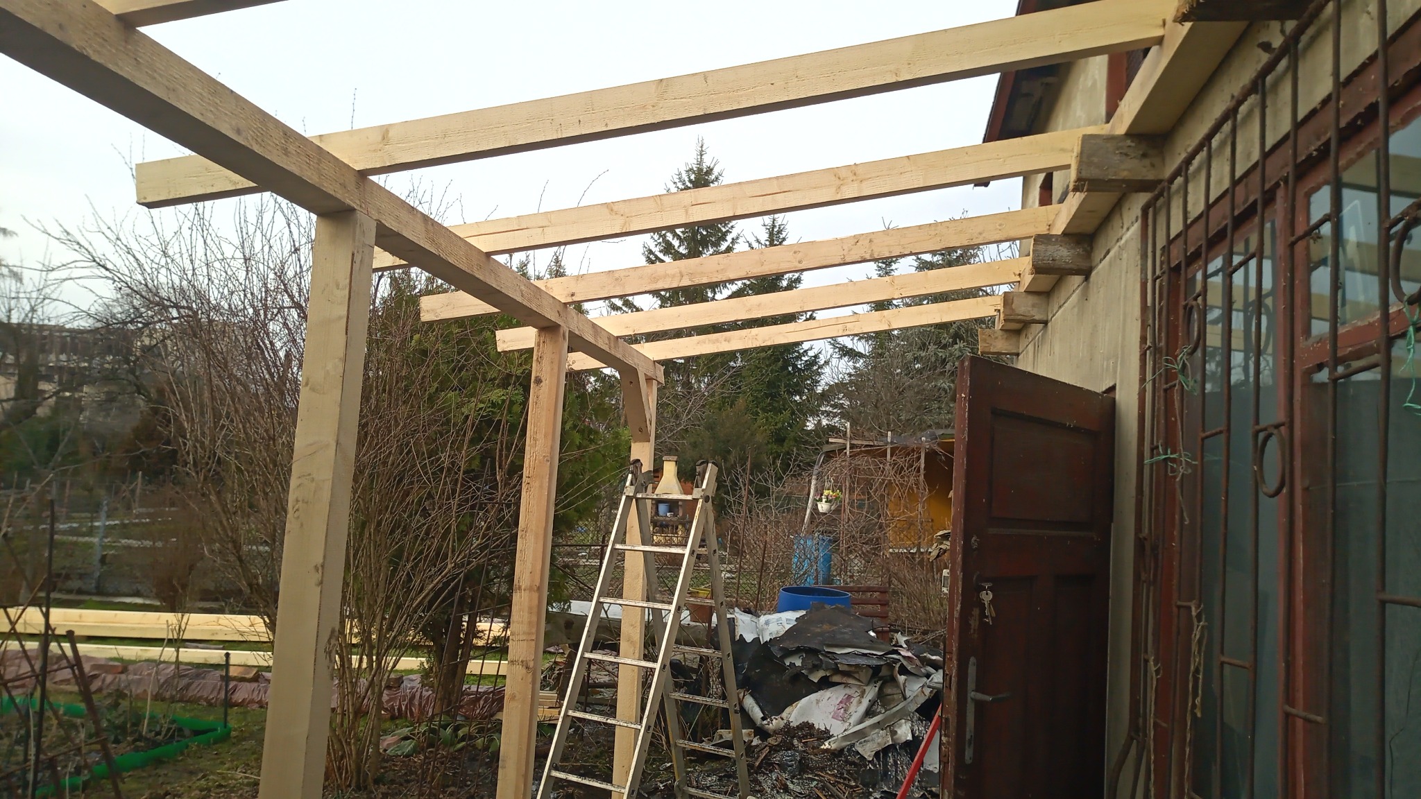 Konstrukcja z surowego drewna, prawdopodobnie zadaszenie tarasu lub werandy, widoczna drabina i materiały budowlane w tle, szara pogoda.