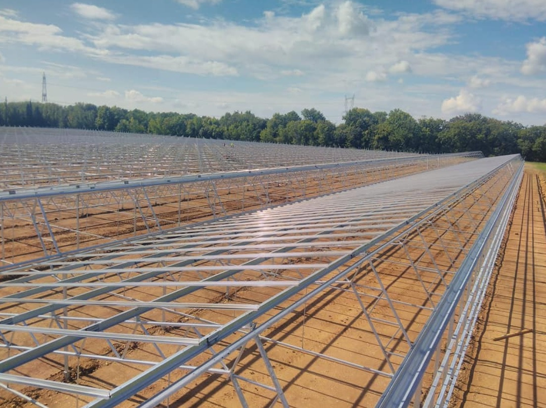 Rozległa konstrukcja stalowa w trakcie budowy, prawdopodobnie pod panele słoneczne, z widoczną linią drzew w tle i błękitnym niebem z chmurami.