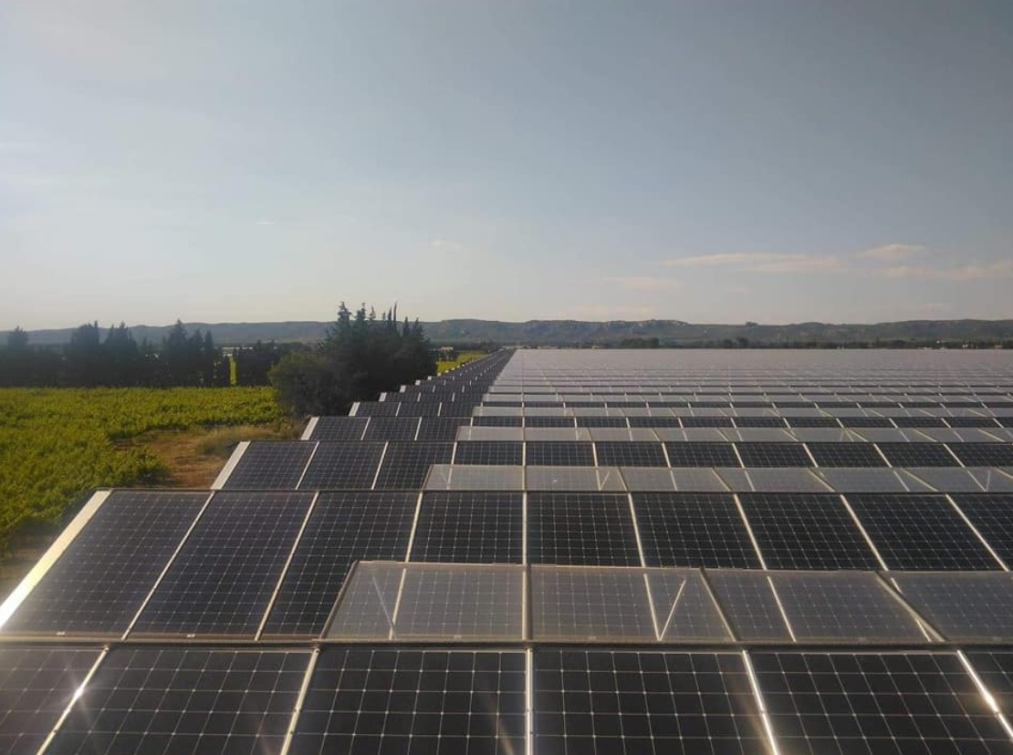 Rozległa instalacja paneli słonecznych na tle zielonych pól i pagórkowatego krajobrazu w oddali, widok z góry.