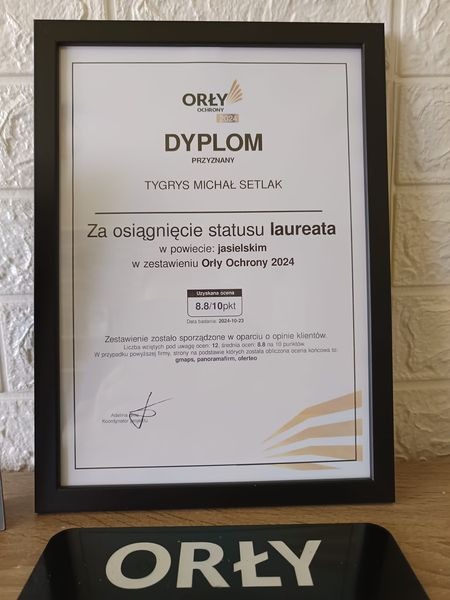 Oprawiony dyplom Orły Ochrony 2024 przyznany firmie Tygrys Michał Setlak za osiągnięcie statusu laureata w powiecie jasielskim, z oceną 8.8/10, widoczny na biurku obok logo Orły.