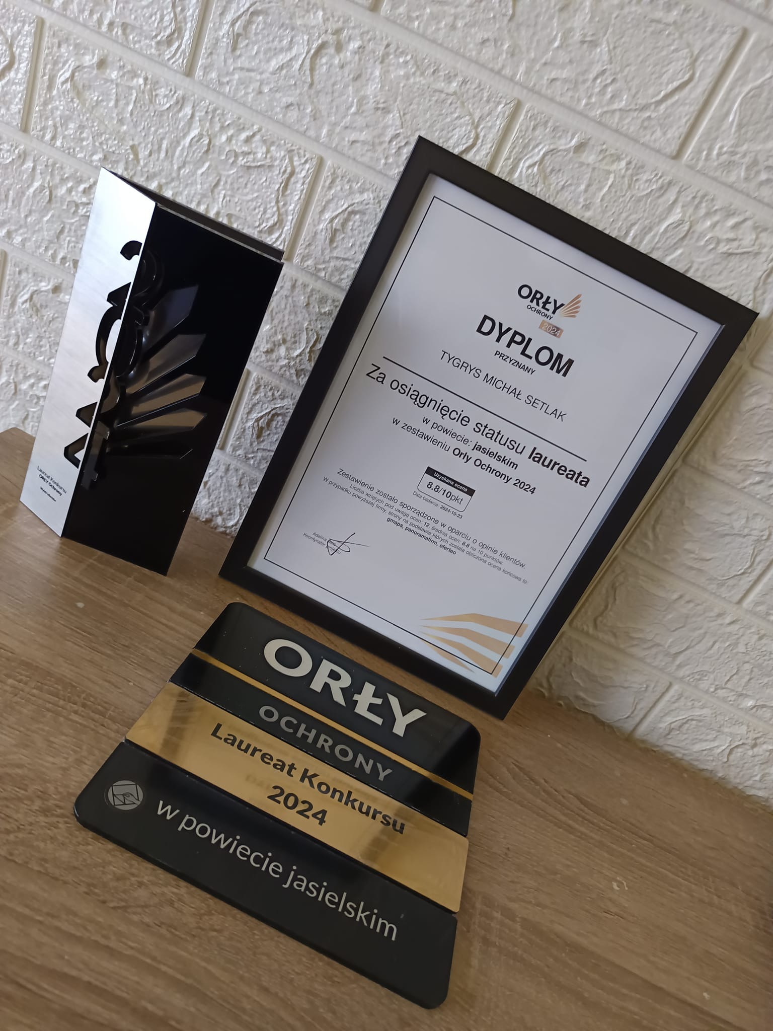 Nagrody i dyplom 'Orły Ochrony 2024' dla firmy ochroniarskiej Tygrys Michał Setlak, obejmujące statuetkę, dyplom z oceną 8.8/10 pkt i tabliczkę laureata konkursu w powiecie jasielskim, na tle...