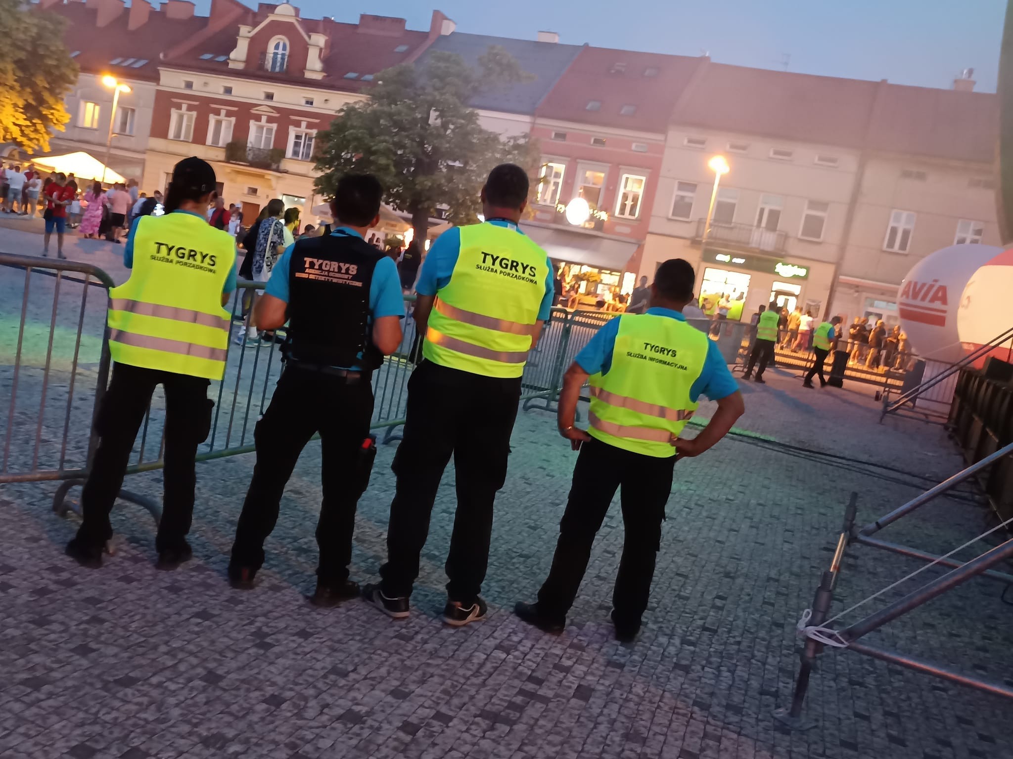 Czterech ochroniarzy firmy 'Tygrys' w kamizelkach odblaskowych i czarnych spodniach, stoi tyłem na placu podczas wieczornego wydarzenia, w tle budynki miejskie.