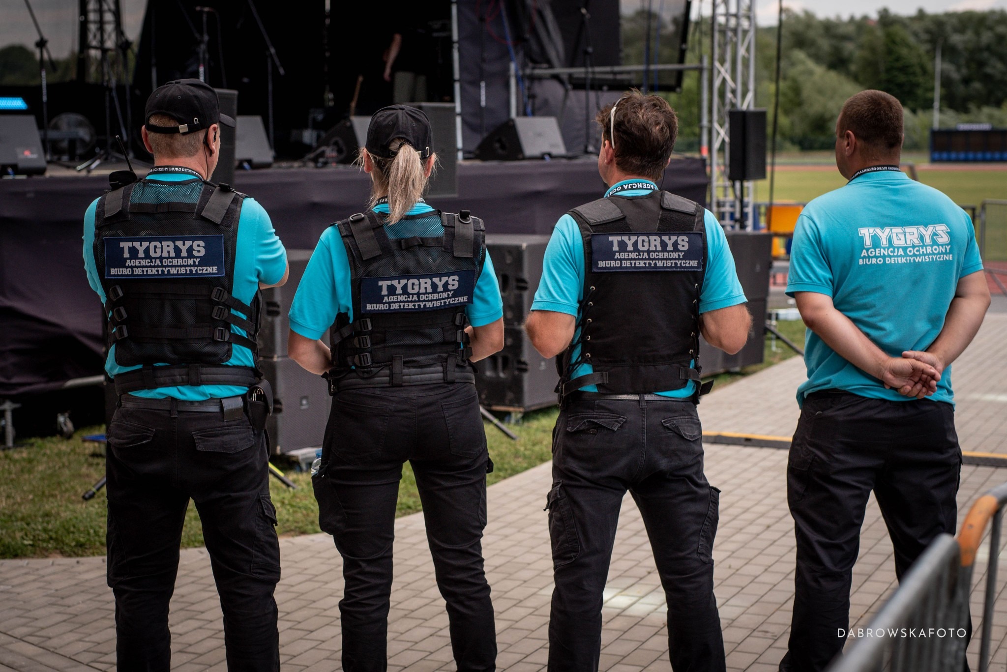 Czterech pracowników ochrony w uniformach z logo firmy 'Tygrys', stojących tyłem na tle sceny i boiska. Widoczne czarne kamizelki i niebieskie koszulki.