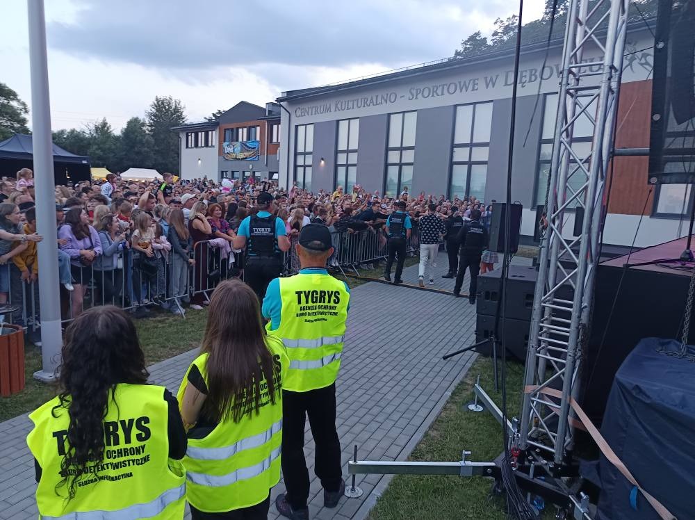 Ochrona imprezy masowej przez pracowników z żółtych kamizelkach z logo. Tłum ludzi za barierkami, budynek Centrum Kultury w tle. Agencja ochrony 'Tygrys'.