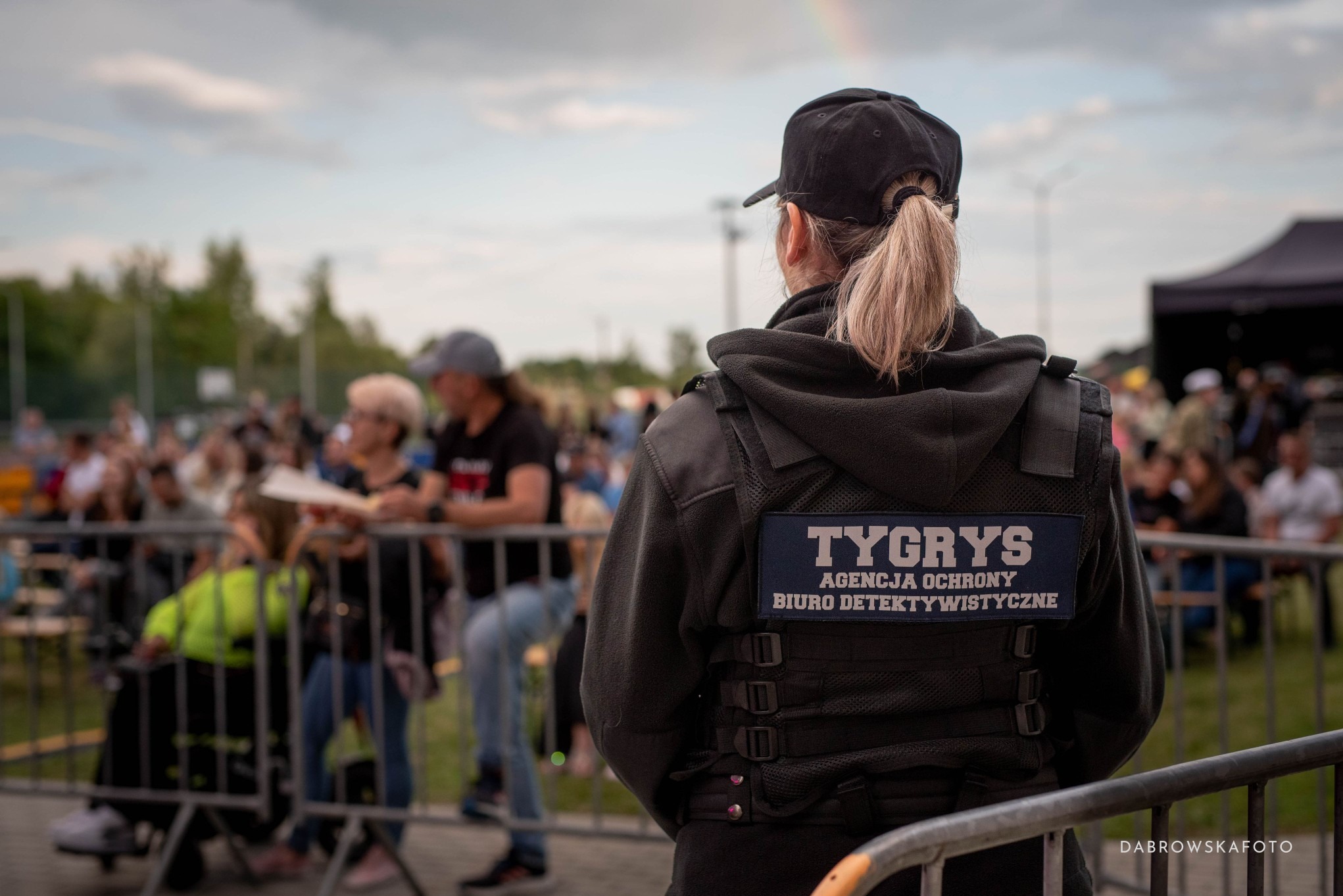 Ochroniarka w kamizelce z napisem 'TYGRYS' na tle tłumu i tęczy na niebie, widziana z tyłu. Zabezpieczenie imprezy masowej przez agencję ochrony.