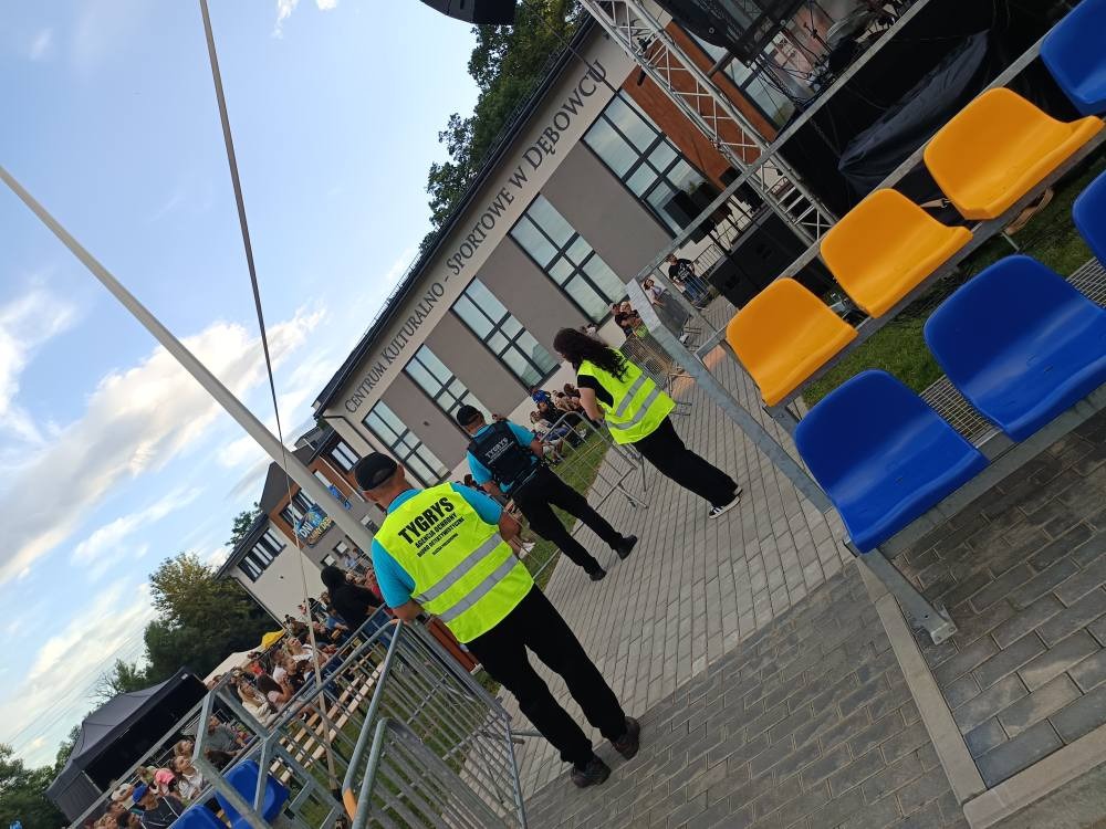 Ochroniarze w żółtych kamizelkach patrolują teren imprezy plenerowej przy Centrum Kultury w Dębowcu. Widoczne trybuny, publiczność i scena w tle. Kąt ujęcia z dołu.