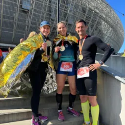 Trzy osoby, mężczyzna i dwie kobiety, stoją na schodach po biegu, owinięci folią termiczną, z medalami na szyjach i numerami startowymi na ubraniach, na tle stadionu.