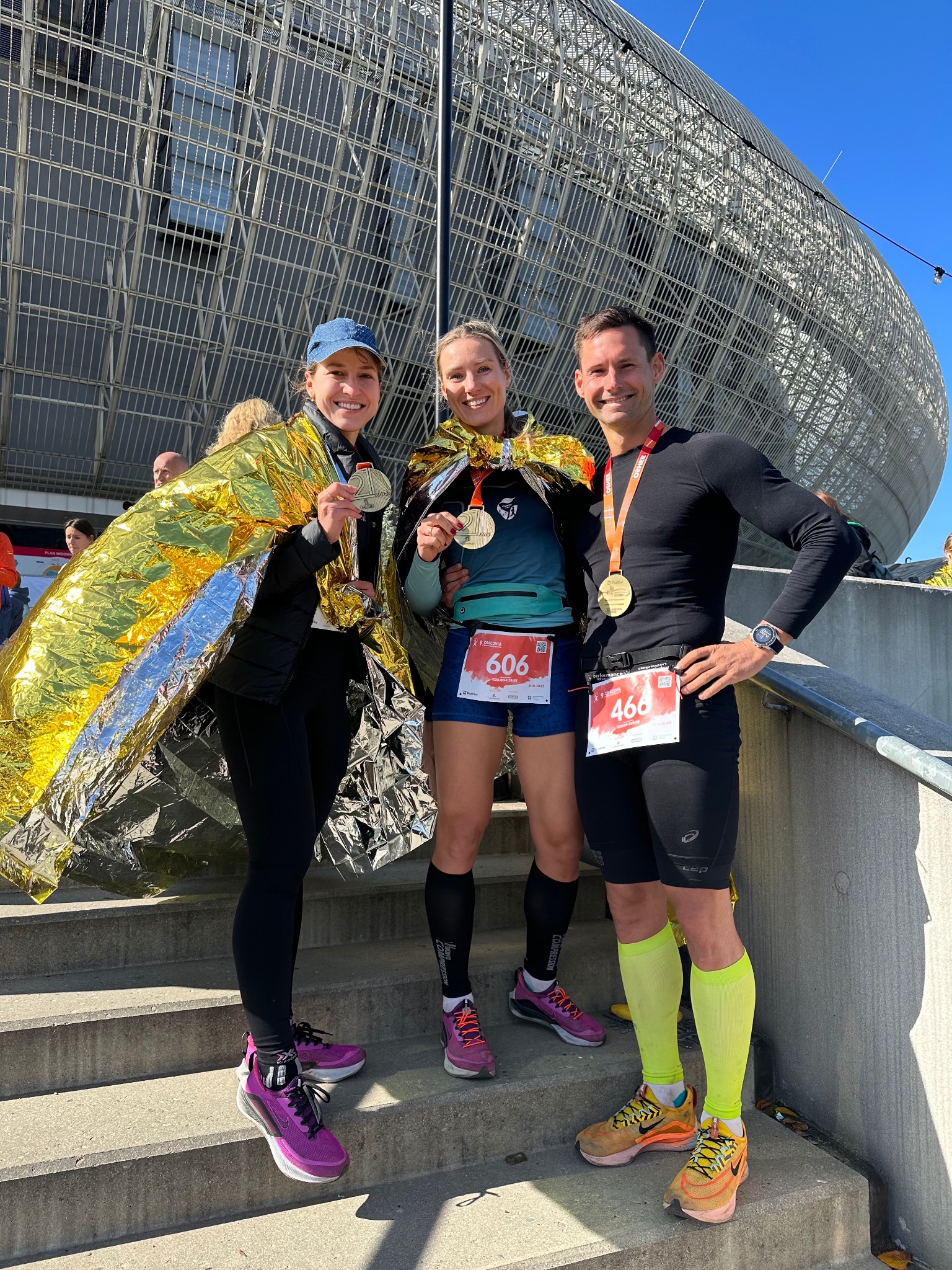 Trzy osoby, mężczyzna i dwie kobiety, stoją na schodach po biegu, owinięci folią termiczną, z medalami na szyjach i numerami startowymi na ubraniach, na tle stadionu.
