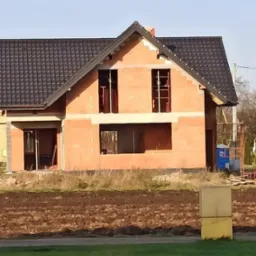 Surowy stan domu z czerwonej cegły z ciemnym dachem, widoczne otwory okienne i drzwiowe, teren wokół budynku przygotowany pod dalsze prace.