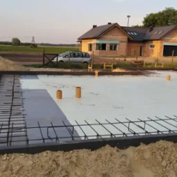 Przygotowany fundament z izolacją i zbrojeniem na tle budowanego domu z pustaków ceramicznych i krajobrazu polnych łąk.