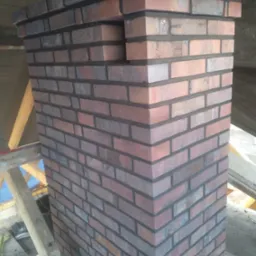 Wymurowany komin z cegły klinkierowej w trakcie budowy, widoczne rusztowanie i elementy konstrukcji dachu.