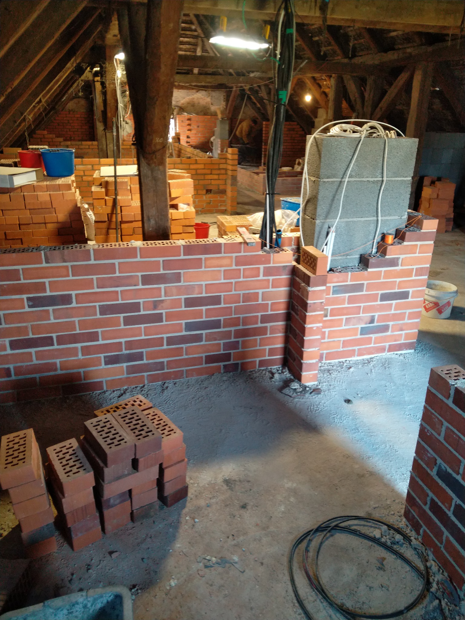 Wnętrze poddasza z częściowo wymurowaną ścianą z czerwonej cegły, widoczne drewniane elementy konstrukcyjne dachu, kable i narzędzia murarskie.