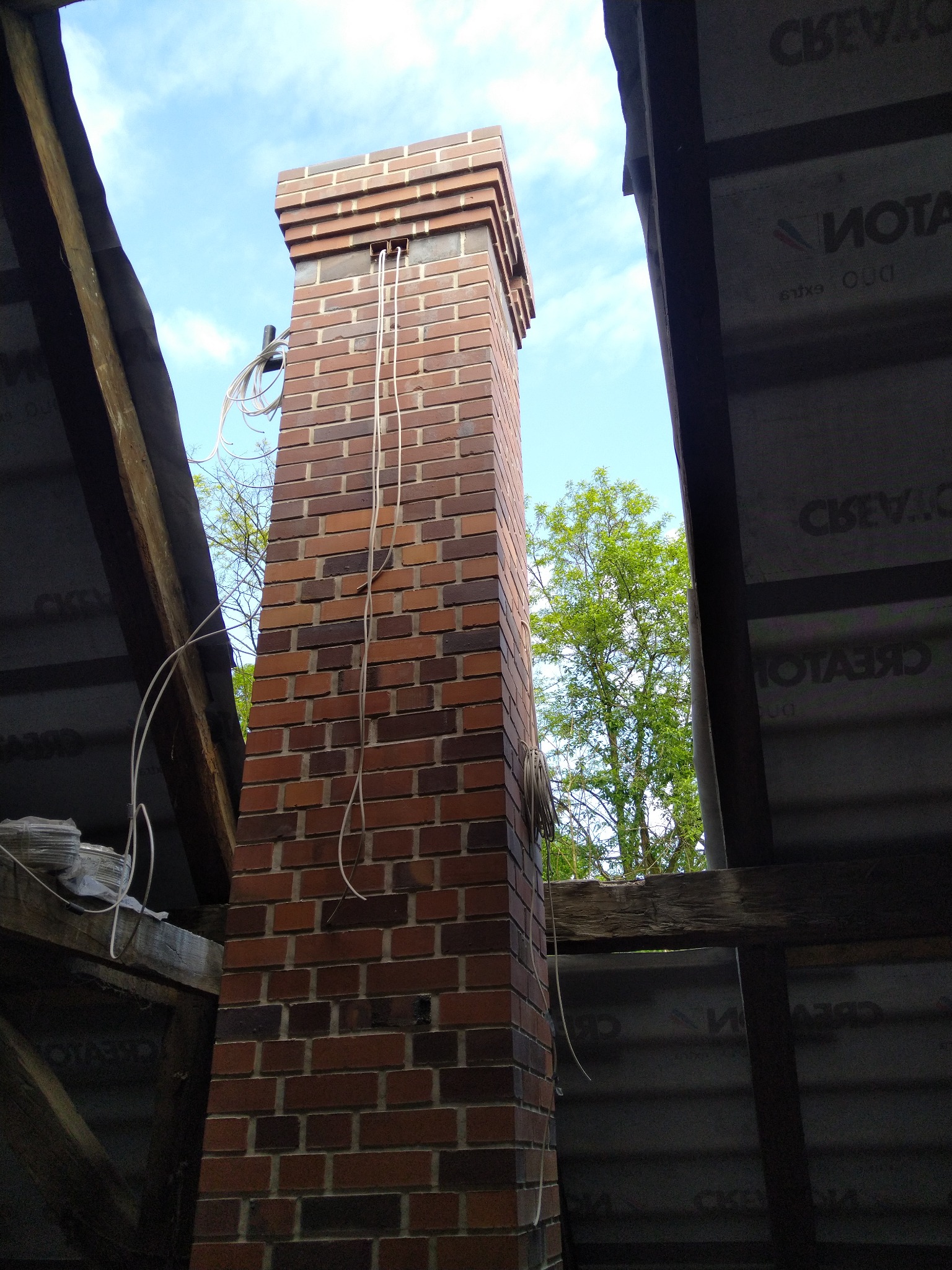 Wewnętrzny widok na murowany komin z czerwonej cegły, wystający ponad konstrukcję dachu, z widocznymi przewodami i fragmentem nieba.
