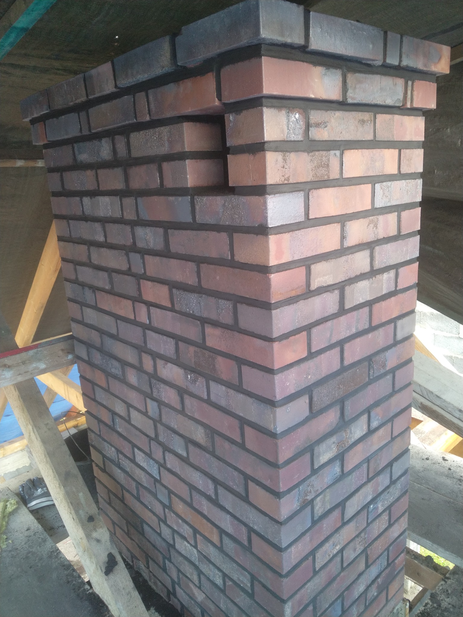 Wymurowany komin z cegły klinkierowej w trakcie budowy, widoczne rusztowanie i elementy konstrukcji dachu.