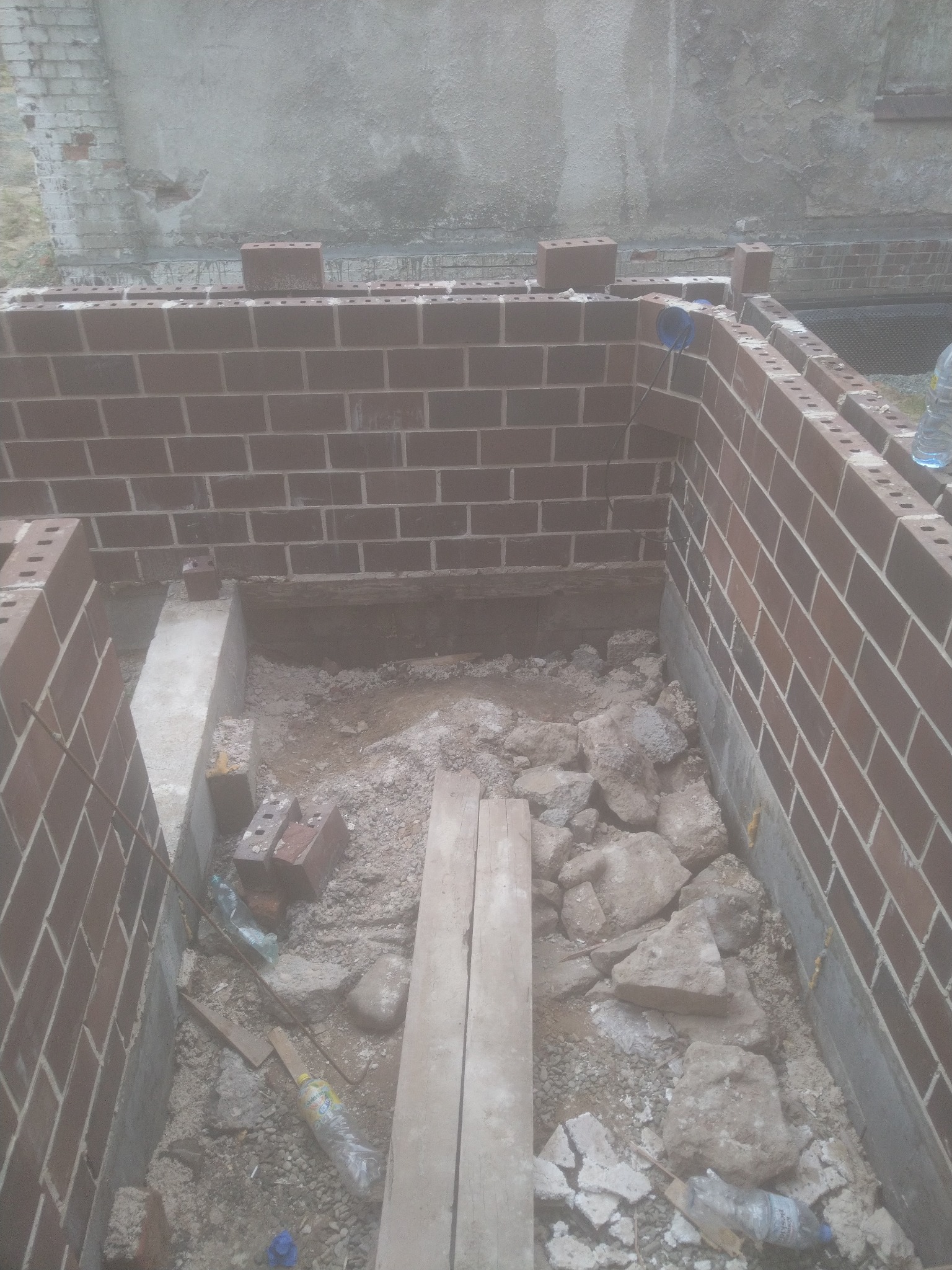 Wykop fundamentowy z częściowo wymurowanymi ścianami z czerwonej cegły, widoczne zbrojenie i gruz na dnie wykopu.