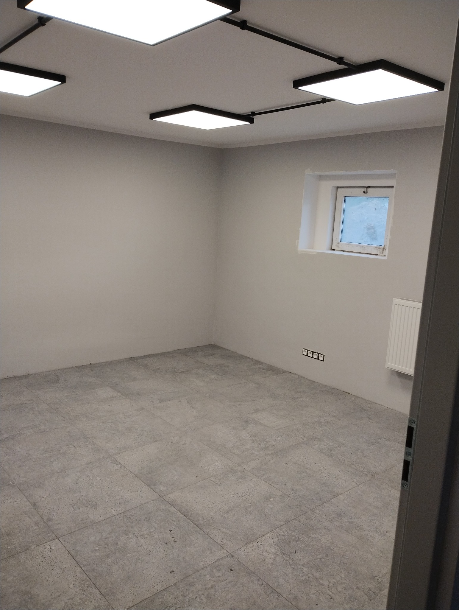 Pusty, szary pokój po remoncie z szarą podłogą z płytek, świeżo pomalowanymi szarymi ścianami, oknem z widokiem na zewnątrz i nowoczesnym oświetleniem sufitowym.