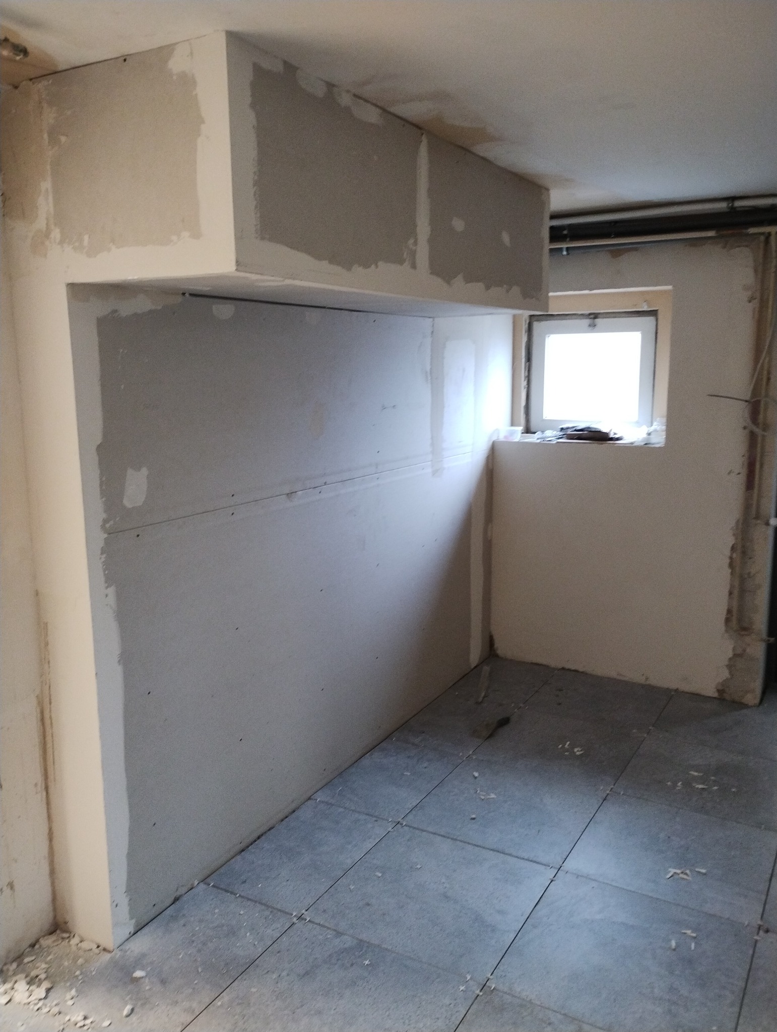 Wnętrze w trakcie remontu: ściany pokryte płytami gipsowo-kartonowymi, sufit z zabudową, szare płytki podłogowe, okno z widocznymi śladami prac budowlanych.
