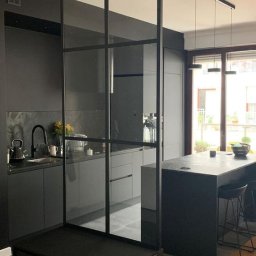 T.wórczo - Nowoczesna, szara kuchnia z wyspą i szklaną ścianką działową z czarnymi szprosami, widok na okno z zielenią na zewnątrz.