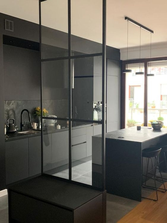 Nowoczesna, szara kuchnia z wyspą i szklaną ścianką działową z czarnymi szprosami, widok na okno z zielenią na zewnątrz.