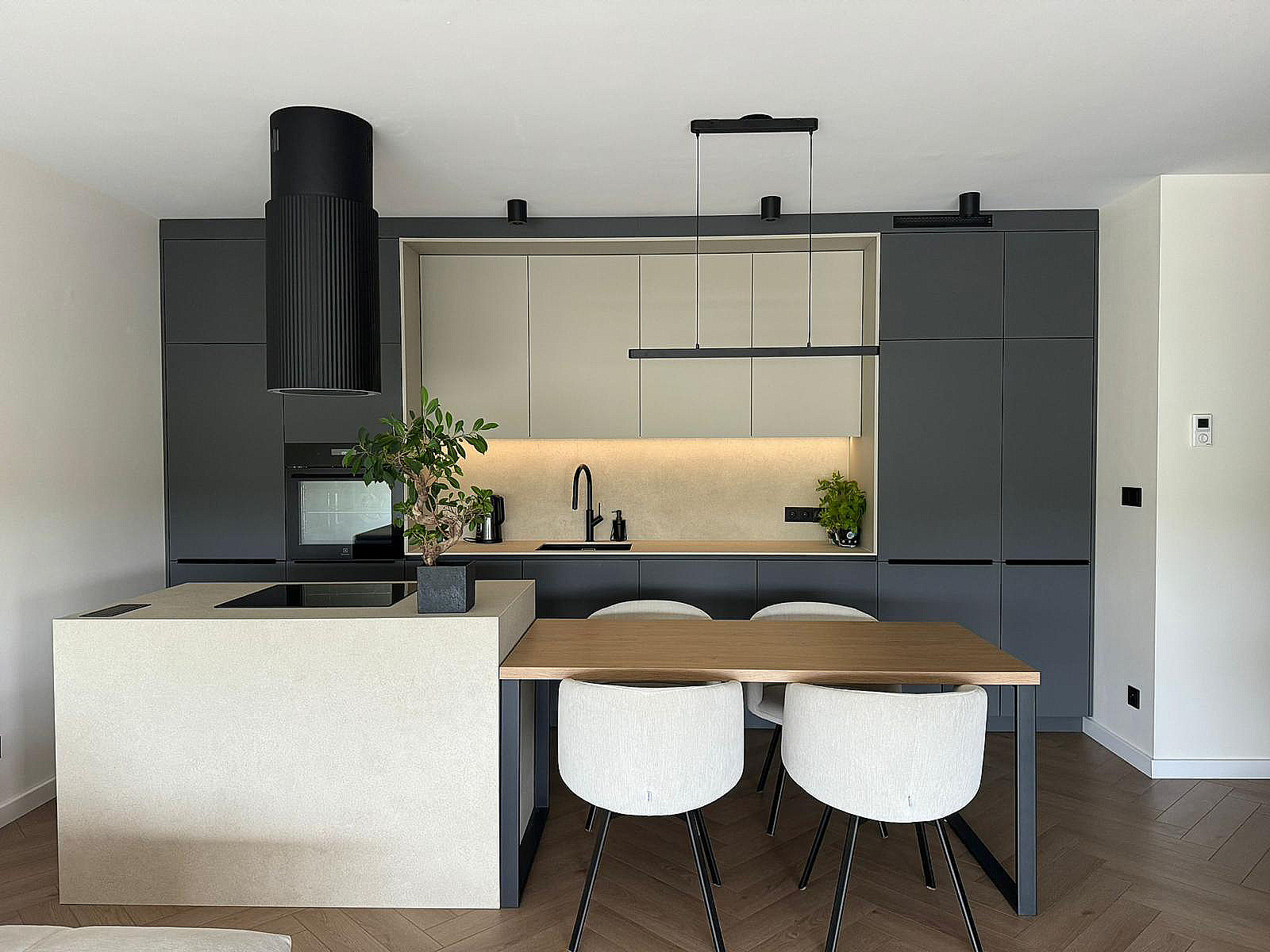 Nowoczesna kuchnia w odcieniach szarości i beżu z drewnianym stołem i białymi krzesłami na parkiecie w jodełkę. Charakterystyczny okrągły okap nad płytą indukcyjną. Oświetlenie LED pod szafkami...