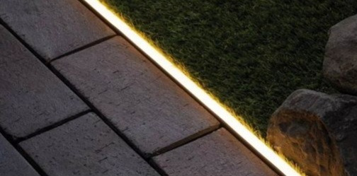 Ścieżka z kostki brukowej oświetlona liniowym, ciepłym światłem LED, przylegająca do trawnika i kamieni dekoracyjnych.