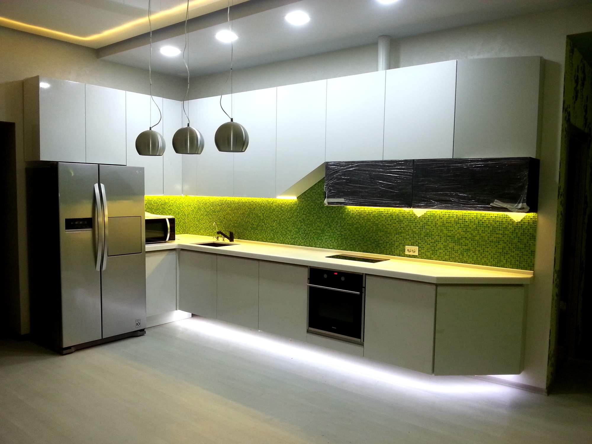 Nowoczesna kuchnia z białymi szafkami, zieloną mozaiką na ścianie, podświetleniem LED pod szafkami i trzema kulistymi lampami wiszącymi.