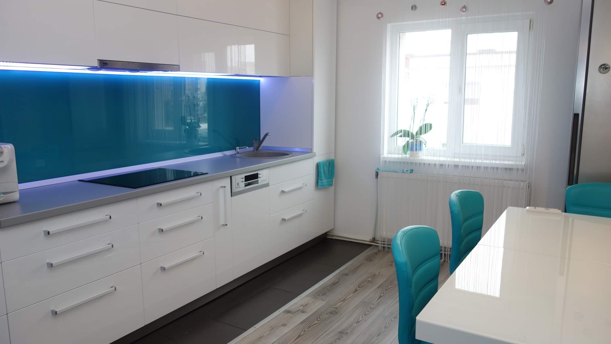 Nowoczesna kuchnia z białymi lakierowanymi szafkami, turkusowym panelem szklanym podświetlonym LED, płytą indukcyjną i turkusowymi krzesłami przy białym stole.