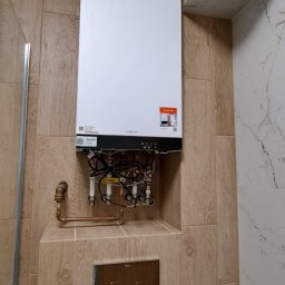 MarGlazur - Nowoczesny piec gazowy Viessmann zamontowany na ścianie wyłożonej płytkami imitującymi drewno w łazience. Widoczne rury i instalacja. Estetyczne wykończenie.